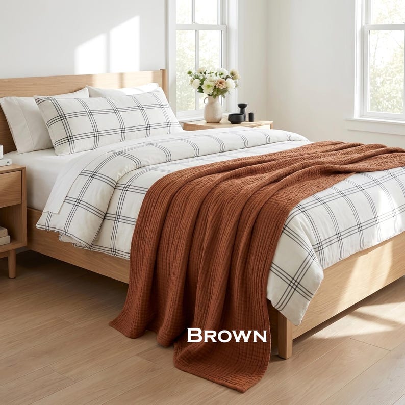 K&ouml;nnte beinhalten: Ein Bett mit einem hellen Holzrahmen, wei&szlig;er Bettw&auml;sche mit schwarzem Karomuster und einer strukturierten, einfarbig braunen Tagesdecke, die &uuml;ber das Fu&szlig;ende drapiert ist. Das Wort BROWN steht in wei&szlig;er Schrift unten in der Bildmitte.