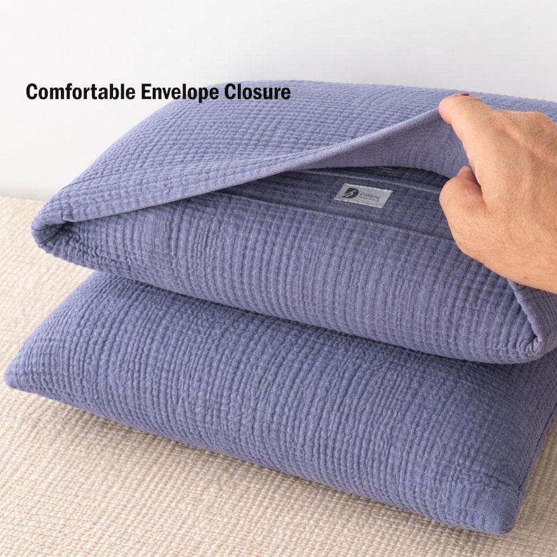 Pode incluir: Duas fronhas empilhadas, na cor azul-arroxeado, com um tecido texturizado e enrugado. A fronha superior est&aacute; parcialmente aberta, revelando o fecho envelope. O texto "Comfortable Envelope Closure" &eacute; vis&iacute;vel.