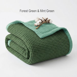 May include: Folded forest green waffle-weave blanket with a mint green border. A mint green top layer is folded over the blanket, and a sprig of cotton bolls rests on top. The text "Forest Green & Mint Green" is above the blanket.