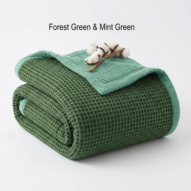 May include: Folded forest green waffle-weave blanket with a mint green border. A mint green top layer is folded over the blanket, and a sprig of cotton bolls rests on top. The text "Forest Green & Mint Green" is above the blanket.