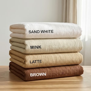 May include: A stack of four folded blankets with a textured, crinkled surface. The blankets are labeled with black text: SAND WHITE for the top, MINK for the second, LATTE for the third, and BROWN for the bottom blanket in the stack.