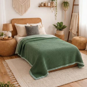 May include: A green waffle-weave blanket with a light blue border is draped over a bed. The bed has a woven headboard and is adorned with pillows. A woven rug and side table with a lamp complete the cozy bedroom setting.