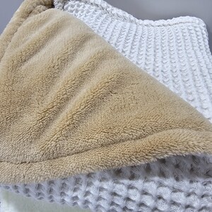 May include: A close-up shot of a beige and white blanket. The blanket features a soft, plush beige interior and a textured white exterior. The white exterior has a waffle-like design. The blanket is folded, showing the contrasting textures and colours.