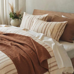 May include: A bed featuring a cream-colored duvet cover and matching pillowcases with thin brown vertical stripes. A solid, textured, rust-colored muslin throw blanket is draped across the bottom. A brown, fuzzy, rectangular pillow sits at the head.