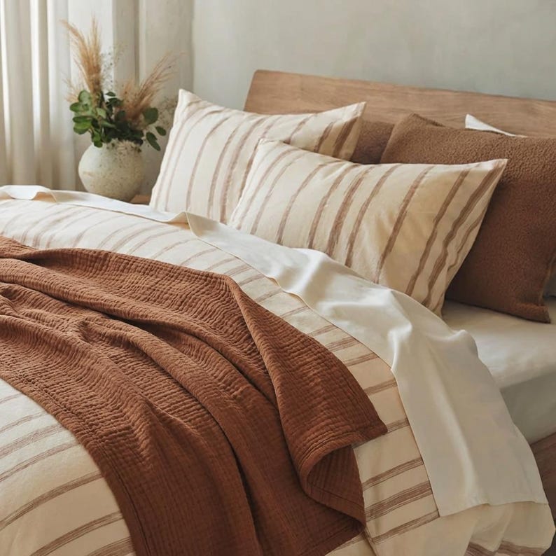 May include: A bed featuring a cream-colored duvet cover and matching pillowcases with thin brown vertical stripes. A solid, textured, rust-colored muslin throw blanket is draped across the bottom. A brown, fuzzy, rectangular pillow sits at the head.