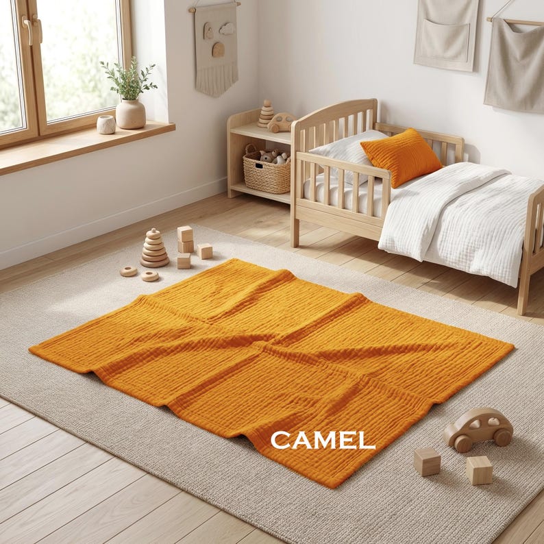 K&ouml;nnte beinhalten: Eine rechteckige senfgelbe Musselindecke mit knittriger Textur und dem in Wei&szlig; gedruckten Wort CAMEL in der unteren rechten Ecke. Die Decke liegt flach auf einem hellen Teppich in einem Raum mit einem Holzbett und verschiedenen Spielzeugen.
