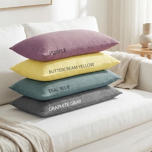 Pode incluir: Quatro almofadas retangulares em tons de roxo, amarelo manteiga, azul-petr&oacute;leo e cinza grafite est&atilde;o empilhadas em um sof&aacute; branco. Cada almofada tem uma superf&iacute;cie texturizada, e os nomes das cores est&atilde;o impressos.