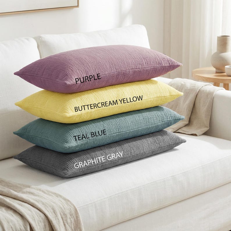 Pode incluir: Quatro almofadas retangulares em tons de roxo, amarelo manteiga, azul-petr&oacute;leo e cinza grafite est&atilde;o empilhadas em um sof&aacute; branco. Cada almofada tem uma superf&iacute;cie texturizada, e os nomes das cores est&atilde;o impressos.