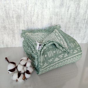 Puede incluir: Una manta de tiro verde y blanca con estampado y borde de flecos. La manta está doblada y tiene una pequeña etiqueta verde con la palabra "Cozy" en ella.