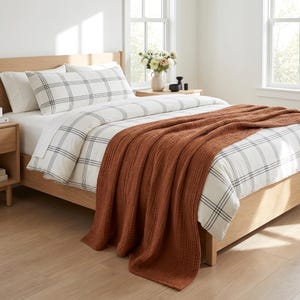 May include: A light oak bed frame features a white duvet cover with a black grid pattern, two matching pillows, and a textured, rust-colored throw blanket draped across the foot of the bed. The bedding set is arranged neatly on the mattress.