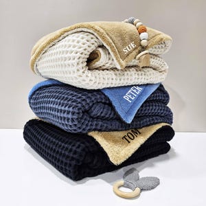 May include: Four stacked waffle-knit baby blankets in cream, navy, and black, with a blue accent. Each blanket is personalised with a name. A wooden and beaded dummy clip and a grey bunny ear teether are included.