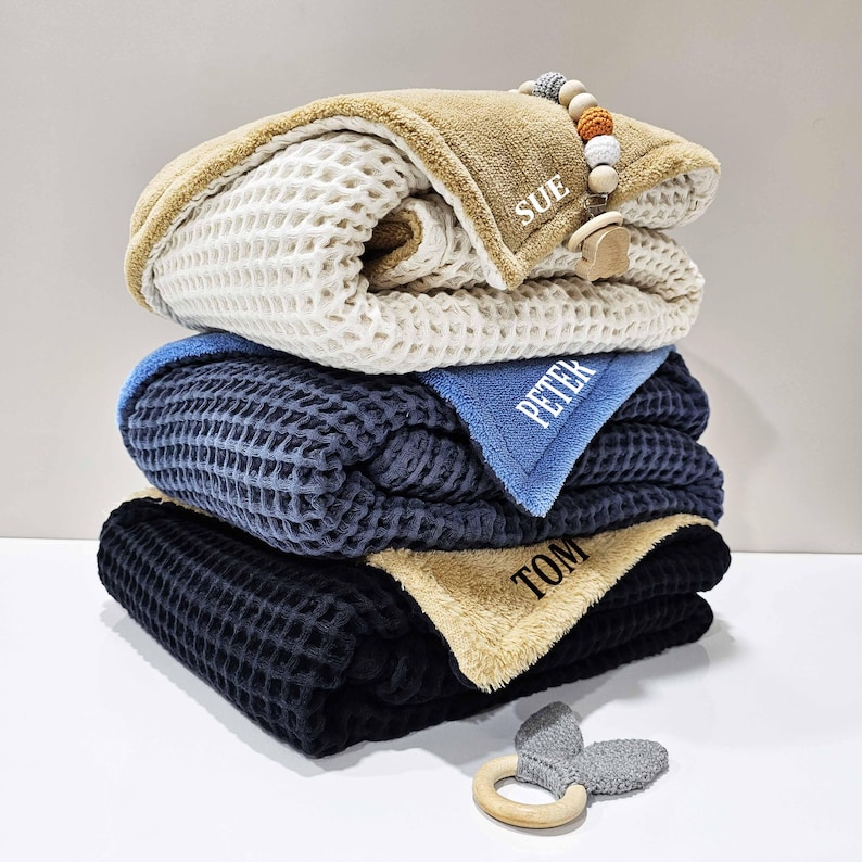 May include: Four stacked waffle-knit baby blankets in cream, navy, and black, with a blue accent. Each blanket is personalised with a name. A wooden and beaded dummy clip and a grey bunny ear teether are included.