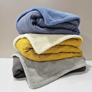 K&ouml;nnte beinhalten: Ein Stapel aus drei gefalteten Decken in Blau-, Gelb- und Graut&ouml;nen. Die oberste Decke ist blau, die mittlere ist gelb mit cremefarbenem Sherpa-Futter und die untere Decke ist grau.