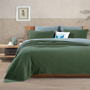 May include: A bed with a green textured blanket and pillows. The bed has a wooden frame and is against a wall with a framed botanical print. Additional pillows are light blue and white with a blue design.