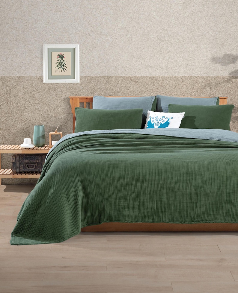 May include: A bed with a green textured blanket and pillows. The bed has a wooden frame and is against a wall with a framed botanical print. Additional pillows are light blue and white with a blue design.
