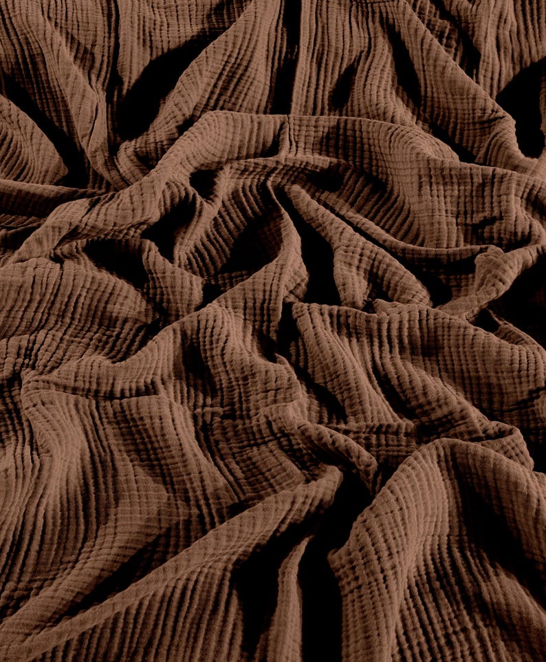 May include: A solid dark brown fabric featuring a distinct crinkled, double gauze texture. The material is draped in deep folds and ripples, creating a pattern of light and shadow across the surface of the soft, lightweight textile.