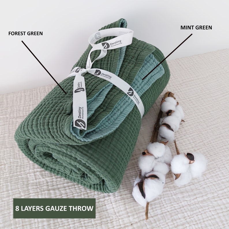 May include: A rolled forest green and mint green 8-layer gauze throw, tied with a white ribbon. The throw is accompanied by a sprig of cotton. The text "8 LAYERS GAUZE THROW" is visible.