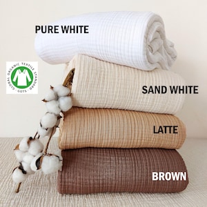 May include: Four different colours of organic cotton muslin fabric, stacked on top of each other. The colours are pure white, sand white, latte, and brown. The fabric is soft and textured, and it is certified by the Global Organic Textile Standard (GOTS).