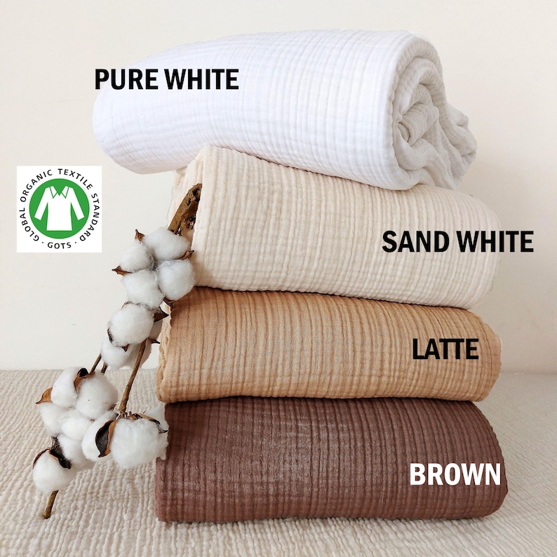 May include: A stack of four folded muslin blankets in pure white, sand white, latte, and brown colors, accompanied by a dried cotton branch. The text labels PURE WHITE, SAND WHITE, LATTE, and BROWN are visible, along with a GOTS organic textile logo.