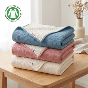May include: A stack of three organic cotton blankets in blue, pink, and cream. Each blanket has a ribbed texture and a contrasting border. The top left corner displays the "Global Organic Textile Standard" logo.