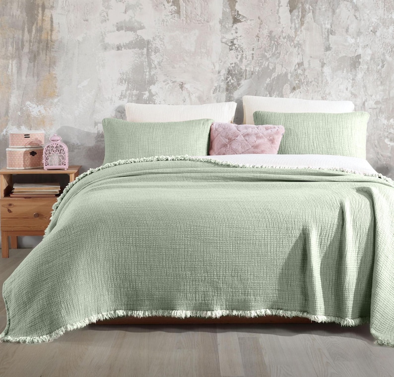 May include: A sage green throw blanket with a fringe border. The blanket is folded on a bed with white and sage green pillows.