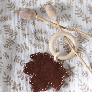 May include: A brown felt leaf with white stitching hangs from a wooden ring. The ring is attached to a string and the leaf is hanging over a white fabric with a brown floral pattern.