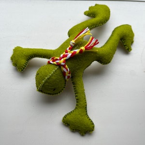 May include: A green felt frog toy with a red, white, and yellow braided scarf. The frog is in a handstand position with its arms and legs outstretched.