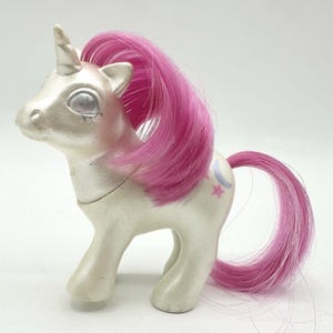 May include: A vintage white unicorn toy with a pink mane and tail. The unicorn has a silver horn and eyes, with a crescent moon and star design on its flank. The toy is made of plastic and is in a standing position.