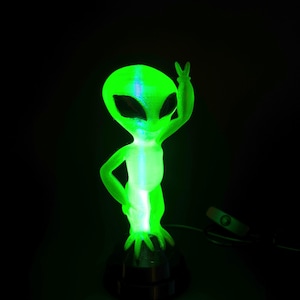 May include: A green alien figurine with a peace sign hand gesture, illuminated from within, creating a glowing effect. The figurine is on a black base and has a cord.