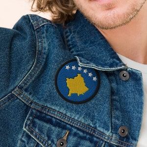 May include: A man wearing a blue denim jacket with a circular patch featuring the flag of Kosovo. The patch has a yellow outline and six white stars.