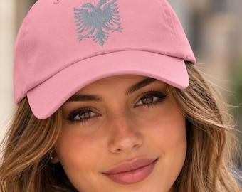 May include: A light pink baseball cap featuring an embroidered grey double-headed eagle emblem on the front panel. The cap has a curved brim, a button on the top crown, and stitched eyelets for ventilation, providing a structured fit.