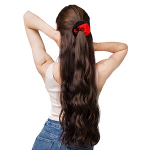 May include: A woman with long, wavy brown hair, wearing a white tank top and blue jeans, has her arms raised behind her head. A black and red scrunchie with a design is in her ponytail. The background is plain white.