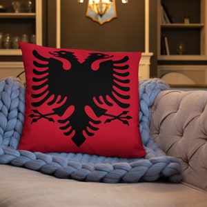 May include: A square red throw pillow featuring a black double-headed eagle design. The pillow is resting on a light gray knitted blanket and a beige sofa. The eagle is a prominent symbol.