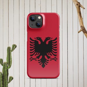 May include: A red smartphone case with a black double-headed eagle emblem. The case is displayed against a backdrop of white wooden planks. The design is a striking representation of a national symbol.