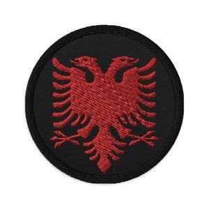 May include: Embroidered patch featuring the Albanian flag's double-headed eagle emblem. The design is rendered in red thread on a black circular background with a black border. The patch is suitable for application on clothing or accessories.