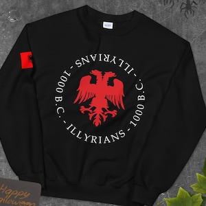 May include: Black sweatshirt with a white circle graphic featuring a red double-headed eagle and the text "ILLYRIANS - 1000 B.C. - ILLYRIANS - 1000 B.C.". A small red flag with a double-headed eagle is visible on the left sleeve.