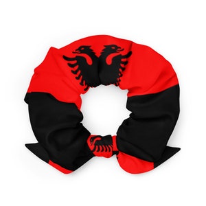 May include: A red and black scrunchie with the Albanian flag design. The flag features a black double-headed eagle on a red background with a black stripe.