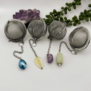 Puede incluir: Cuatro infusores de té de plata con cadenas y abalorios de piedras preciosas. Los abalorios son una lágrima azul, una hoja amarilla, un cubo morado y un rectángulo verde.
