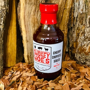 May include: A glass bottle of Smokey Moe's Cherry Chipotle Sauce with a red lid. The bottle is sitting on a bed of wood chips. The label says "Smokey Moe's" and "Cherry Chipotle Sauce".