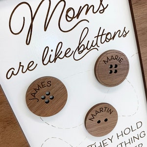 May include: A framed wooden sign with the text "Moms are like buttons, they hold everything together". The sign has three wooden buttons with the names "James", "Marie", and "Martin" engraved on them.