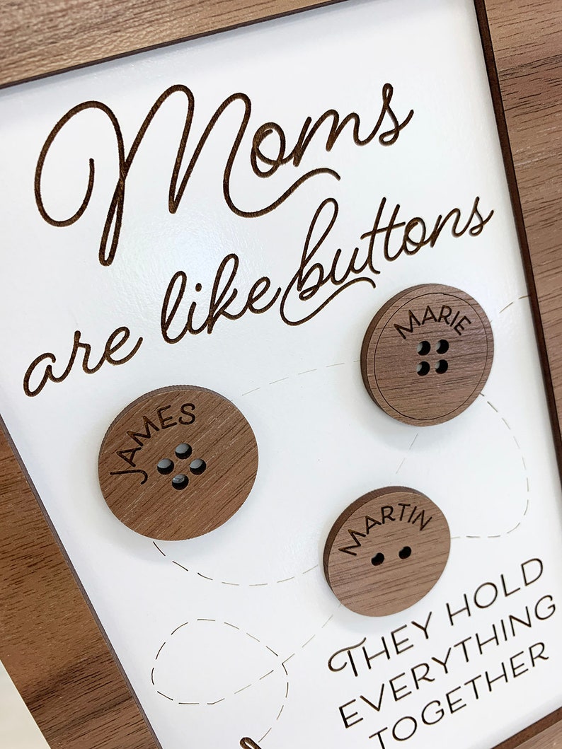 May include: A framed wooden sign with the text "Moms are like buttons, they hold everything together". The sign has three wooden buttons with the names "James", "Marie", and "Martin" engraved on them.
