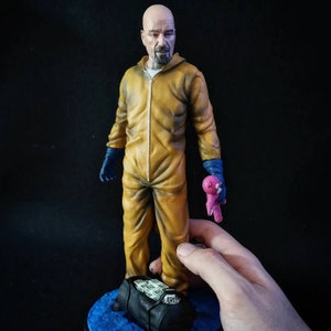 May include: A yellow jumpsuit figurine of a bald man with a beard, holding a pink teddy bear. The figurine is standing on a blue base with a black bag filled with money.