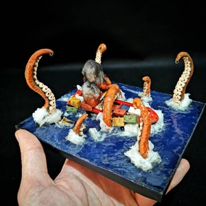 May include: A miniature model of a cargo ship being attacked by a giant octopus with multiple tentacles. The ship is surrounded by containers and the octopus is made of orange, white, and black materials. The scene is set on a blue surface with white foam.