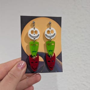 Könnte beinhalten: Ohrringe zum Aufhängen mit einem geschichteten Design. Jeder Ohrring hat einen weißen Totenkopf, eine grüne Maske und eine rote Maske. Die Ohrringe sind an einem silberfarbenen Haken befestigt. Sie werden auf einer Karte mit gelbem Mondhintergrund präsentiert.
