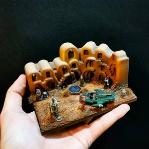 May include: A miniature desert scene with a small, green spaceship, a blue pool, and several small figures. The scene is built on a brown base with sand-colored details.