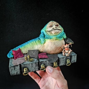 May include: A hand-painted figurine of Jabba the Hutt, a large, slug-like alien from Star Wars, reclining on a platform with a smaller alien figure beside him. The platform is decorated with pillows and other details.