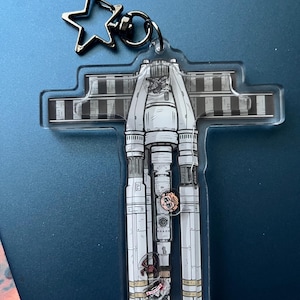 May include: Clear acrylic keychain featuring a detailed illustration of a spaceship design in white, gray, and black. The keychain has a silver star-shaped charm and a silver keyring. The design includes intricate details of the spaceship's structure.