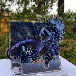 May include: A layered acrylic display featuring a detailed illustration of a blue alien creature with sharp claws and teeth. The artwork includes a background with a dark, ominous atmosphere and a smaller alien figure. The display stands on a clear base.