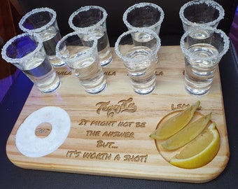 May include: A wooden tequila shot board with eight shot glasses filled with clear liquid, a small dish of salt, and three lemon wedges. The board has the text "Tequila. It might not be the answer but... It's worth a shot!" 