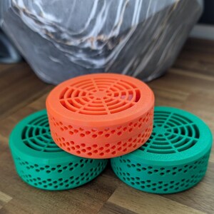 May include: Three circular, vented objects in vibrant colours: orange and two shades of green. Each has a circular top with a fan-like design and a perforated side. The objects are stacked on top of each other.
